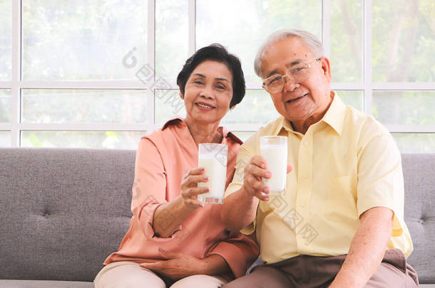 健康的老年人生活方式概念。健康的亚洲老年夫妇坐在客厅的沙发上，举着奶杯，在镜头前表演.