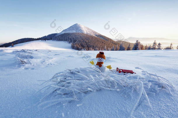 戴着帽子、手套和围巾的雪人拉雪橇.美丽的冬日新年快乐。草坪上覆盖着雪.高山的风景。新年快乐.