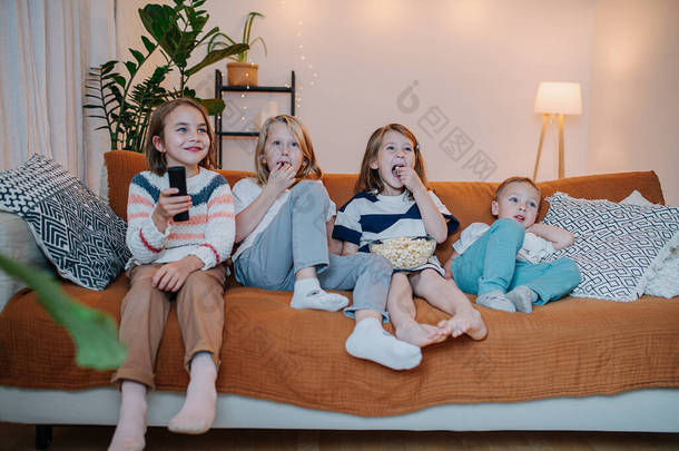 一组5岁至10岁的儿童坐在沙发上看电视，吃爆米花