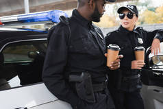 多文化警察，戴着纸杯，靠着巡逻车在室外模糊的背景下交谈