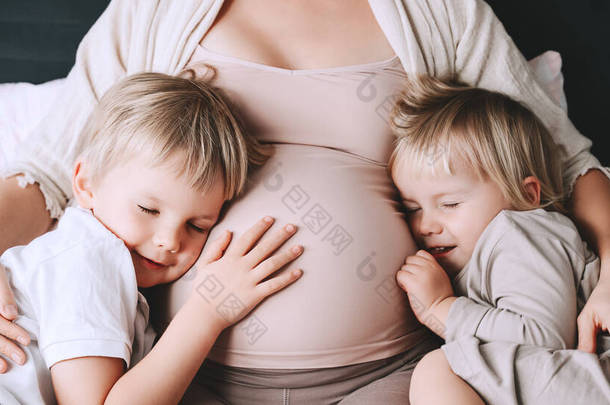 怀孕妇女和她的孩子在床上休息。可爱的母亲和幼儿一起呆在家里。小孩子们牵着妈妈怀孕的肚子。第三次怀孕母亲、家庭、养育子女的概念.