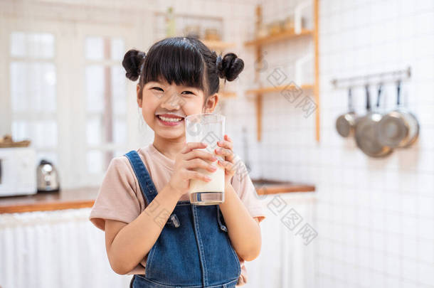 在家里厨房里拿着一杯牛奶的亚洲<strong>可爱</strong>小孩的画像。学龄前小女孩或女儿笑着<strong>呆</strong>在家里，开心地喝牛奶，然后看着相机.