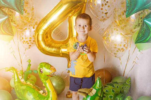 在一个装饰着金黄色、绿色和黄色气球的生日宴会的房间里，有一个小男孩，巨大的充气4号，他带着盒子和缎带。照片拍摄区域在家里。恐龙狒狒.