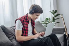 集中的青少年与耳机玩游戏的笔记本电脑, 而坐在沙发上