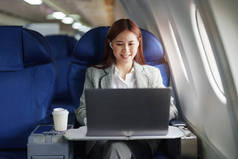 一位成功的亚洲女商人或穿着正装的女企业家坐在商务舱座位上，在飞行过程中使用电脑膝上型计算机.