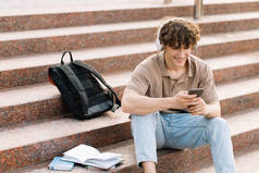 迷人的卷曲头发的年轻人大学生或大学生坐在楼梯上，看着智能手机