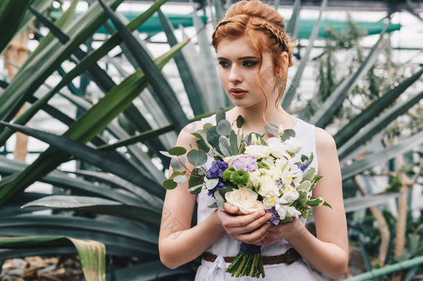 美丽的年轻红发新娘举行婚礼花束和看着远离植物园 