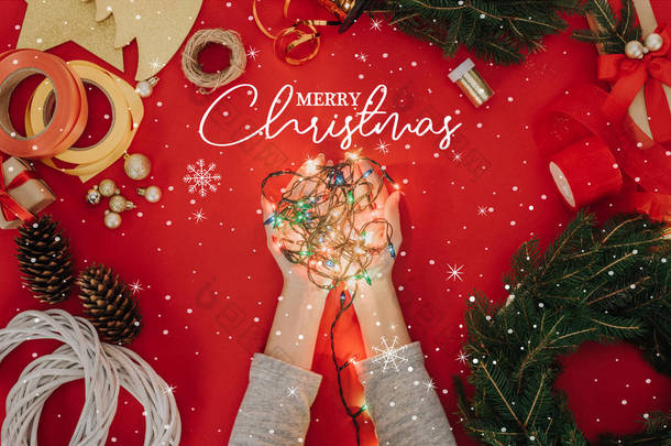 裁剪拍摄的妇女拿着<strong>圣诞</strong>树的灯在手与松树树枝和装饰手工花圈在红色的背景下, 用 