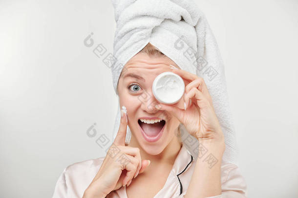 兴奋的女孩，头戴毛巾，左眼拿着一罐面霜涂在脸颊上