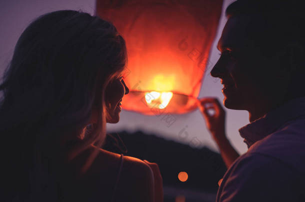 一对夫妇的肖像在晚上在河海滩上发射天空灯笼, 看着对方