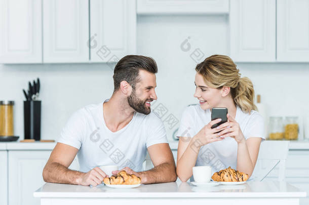 快乐的年轻夫妇互相<strong>微笑</strong>, 并使用智能手机, 同时共进早餐