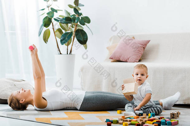 迷人的女人躺在地毯上, 做运动与哑铃和可爱的幼儿男孩拿着木箱在客厅