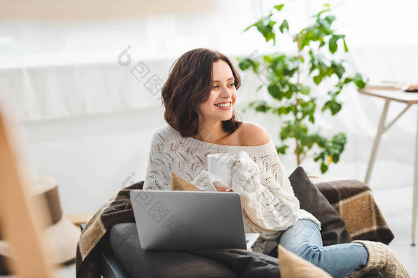 在舒适的家里用笔记本电脑喝茶的漂亮而快乐的女人