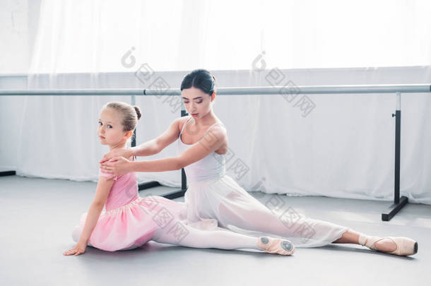 可爱的小芭蕾舞演员在粉红色的芭蕾舞短裙看着相机, 而与教师在芭蕾学校锻炼