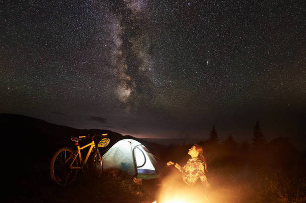 年轻女子游客在夜间露营, 在燃烧的篝火旁, 照亮的旅游帐篷, 山地自行车在惊人的美丽的夜空充满星星和银河。天文<strong>摄影</strong>