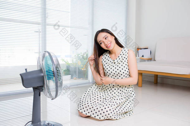 美丽的亚洲妇女坐在客厅地板上享受电风扇冷风.