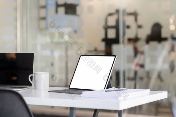 用空白屏幕平板电脑、笔记本电脑、办公用品和咖啡杯在有模糊办公室的白桌子上裁剪的工作场所照片 