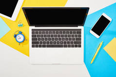 笔记本电脑、智能手机、数字平板电脑的顶视图，带空白屏幕，背景为黄色、蓝色和白色，配有办公用品