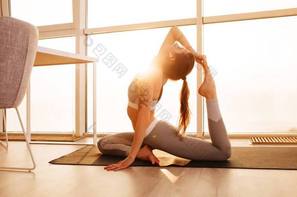 年轻漂亮的女士在运动的顶部和绑腿的肖像和训练<strong>瑜伽</strong>体式<strong>瑜伽垫</strong>在家里与大窗口背景