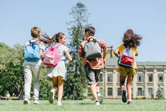 四个多民族学童背着背包在公园的草坪上奔跑的背景