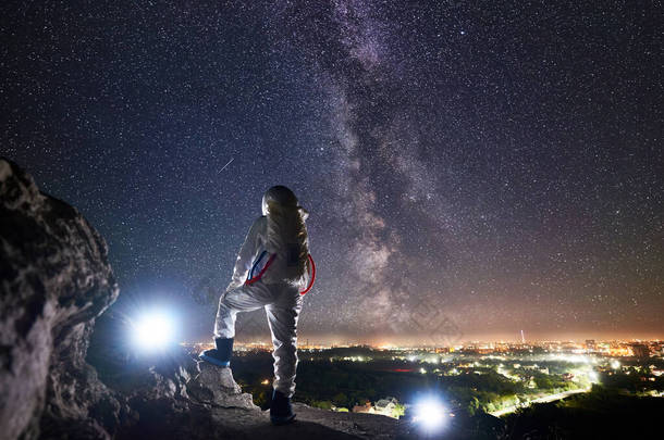 可以看到太空旅行者站在岩石山上，<strong>欣赏</strong>着星空、银河和夜城的美景。身穿白色宇航服的飞行任务专业宇航员。空间旅行的概念