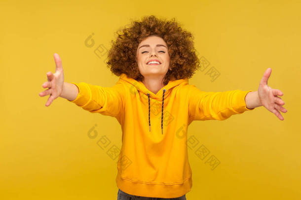 到<strong>我</strong>怀里来！一个极其快乐、友善、卷发、穿着都市风格帽衫的女人的画像，伸出双手拥抱，给予自由的拥抱和欢迎。室内拍摄，黄色背景