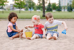 三可爱的白种人和拉美裔儿童的肖像, 孩子们坐在沙盒里玩塑料五颜六色的玩具。小女孩和男孩朋友一起在操场上玩得开心. 