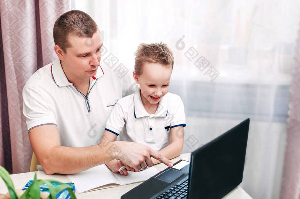 家庭远程在线学习。爸爸和儿子坐在窗边的桌子旁，在笔记本电脑上看视频课。文字的位置。检疫验尸官。父亲节.