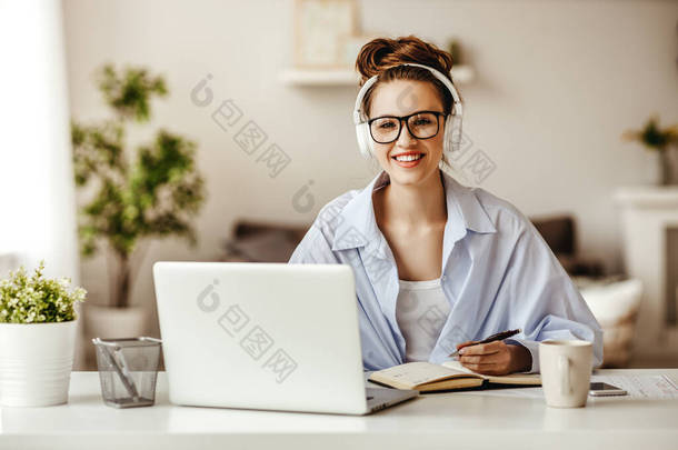 专心致志的女自由职业者，戴着眼镜和无线耳机，一边对着相机微笑，一边坐在笔记本电脑旁，一边喝着一大杯热饮，一边用记事本写着轻松的现代生活