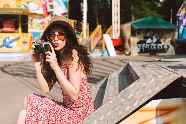 美丽的女士与深色卷曲的头发在太阳镜和帽子坐在手里的小相机和花时间在游乐园与景点<strong>背景</strong>