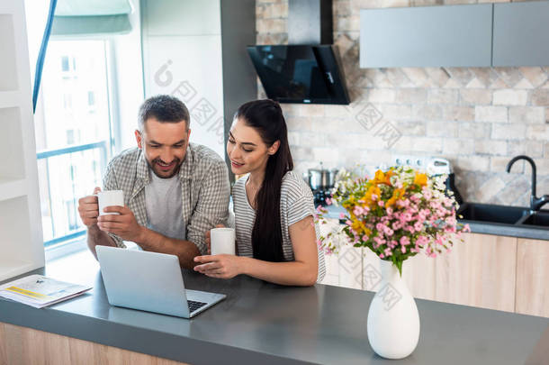 微笑的已婚夫妇看笔记本电脑屏幕一起在柜台在厨房