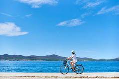 女孩骑单车在白色夏天关闭和帽子骑自行车沿石人行道上蓝色波光粼粼的海水和度假村在山脚下的对岸背景。旅游和度假.