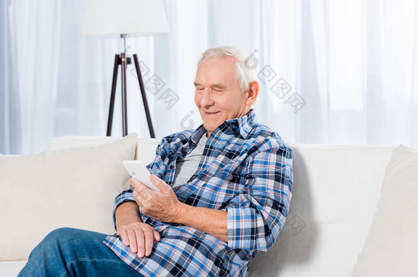 坐在沙发上, 在家里使用智能手机的微笑老人的肖像
