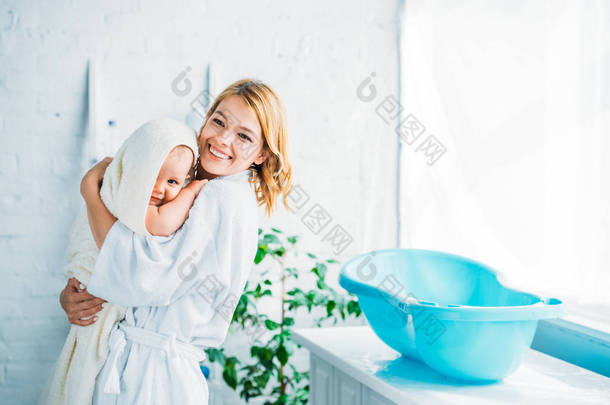 快乐的母亲在浴袍携带可爱的孩子覆盖在毛巾附近塑料婴儿浴缸