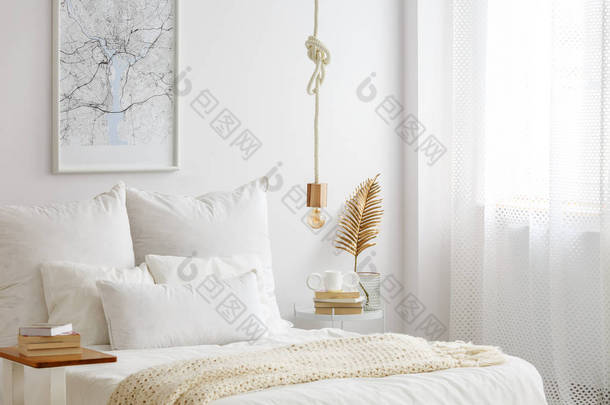 在<strong>床</strong>上挂着白色被褥、书和金色蕨叶的绳子上的简单灯泡灯在白色卧室内部的一个末端桌上