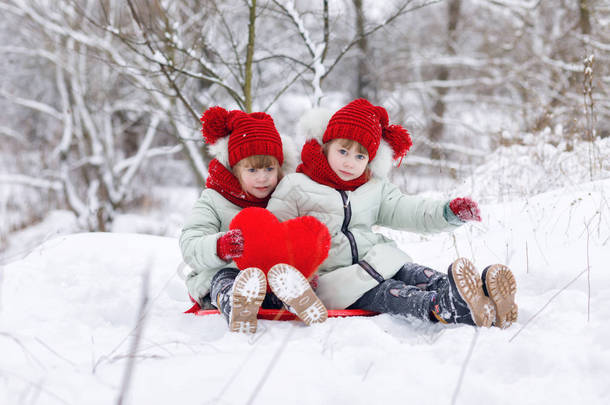 有趣的姐妹双胞胎坐在雪地上, 在冬季公园散步时, 手里拿着一颗巨大的红心.