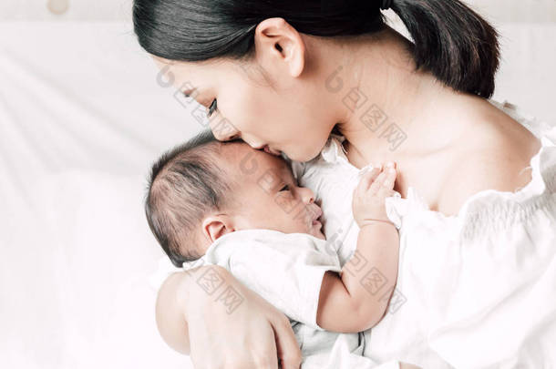 母亲抱着<strong>婴儿</strong>在她的怀里, 亲吻在一个白色的卧室。对家庭概念的爱