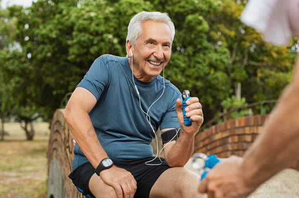 快乐的老人听音乐在耳机和行使使用手爪。微笑的老人做运动, 而看他的妻子在公园。老人训练加强手的肌肉.
