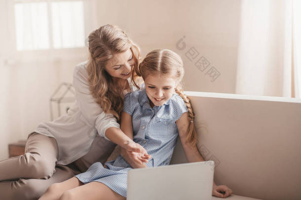 幸福的母亲和女儿坐在沙发上和在家里使用笔记本电脑