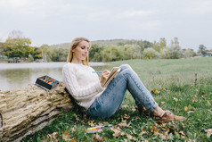 穿着白色毛衣和蓝色牛仔裤的漂亮微笑的年轻女子画在笔记本上，彩色铅笔坐在草地上，靠在湖面上的一棵干树的树干上