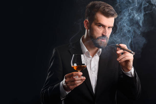 抽烟的男人喝酒。穿着正式服装的有吸引力的商人的横向<strong>肖像</strong>。一杯白兰地和一支烟熏雪茄在他手中.
