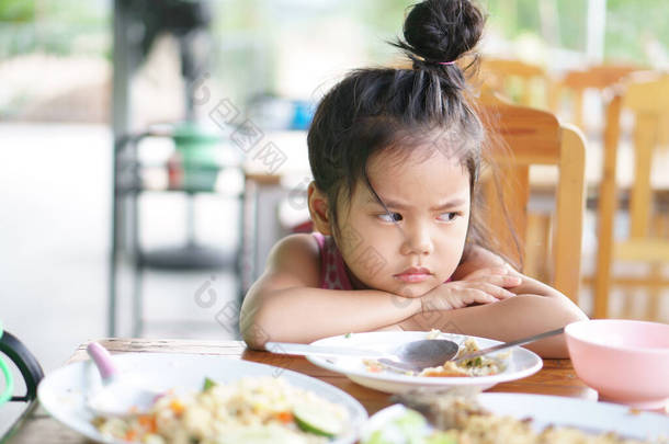亚洲小孩可爱或小孩厌食症或伤心无聊的食物或困倦的空余食物撑着身子或面颊靠在木桌上吃早餐或午餐