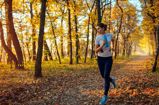 跑步者在秋季公园里锻炼。 日落时拿着水瓶跑的女人. 积极健康的生活方式