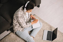 坐在笔记本电脑附近的地板上,在笔记本上写字的头戴耳机的年轻人的头顶视图