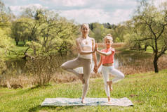 运动妈妈和女儿一起练习瑜伽在新鲜空气中，Vrikshasana瑜伽姿势或树的姿势。健康的生活方式、运动、健康