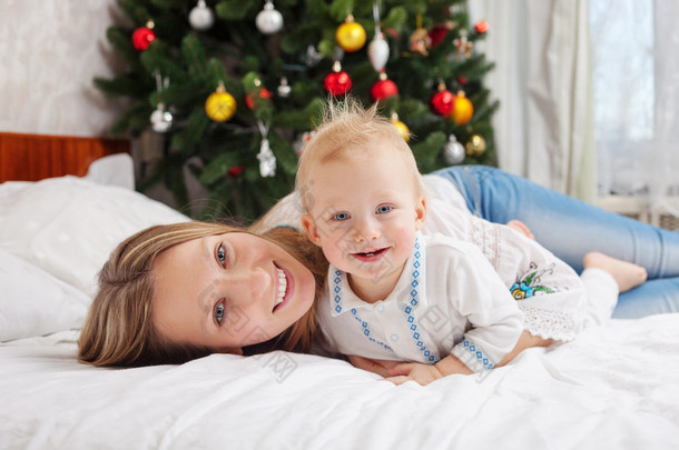 快乐妈妈和宝贝男孩在床上在家里与背景装饰圣诞树的肖像