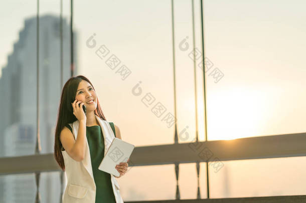 美丽年轻的亚洲女商人使用智能手机和数字平板看向上到副本空间，商务沟通或移动会议概念，现代办公背景日落与 rim 光
