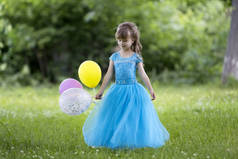 漂亮的金发长发女孩在漂亮的长蓝色晚礼服举行彩色气球站在盛开的田野上, 在晴朗的阳光明媚的日子, 在模糊的绿树背景。快乐童年概念.