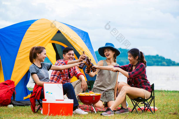 组的男人和女人享受露营野餐和烧烤在帐篷中<strong>背景</strong>的湖。年轻混合种族亚洲女人和男人。年轻人们手里烘培和欢呼瓶啤酒.