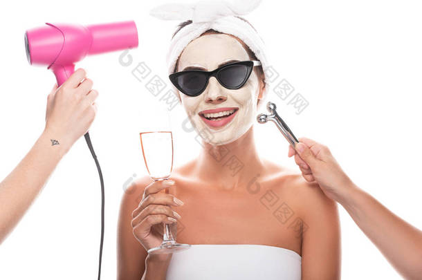 部分视图美容师拿着吹风机和面部按摩器和微笑的女人在太阳镜与面部面具拿着酒杯隔离在白色